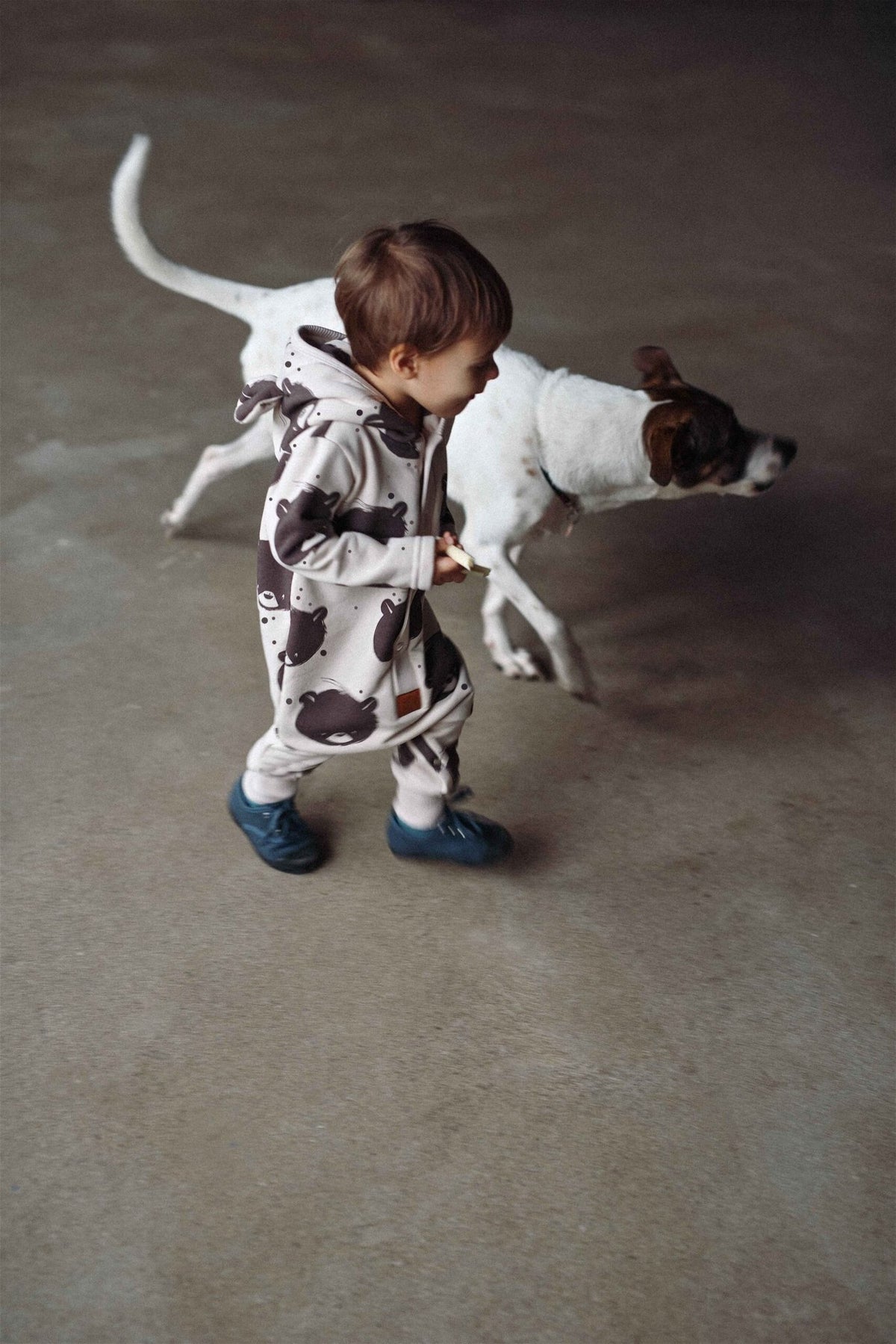 1. Child in beige bear-patterned jumpsuit with hood running alongside a dog indoors
