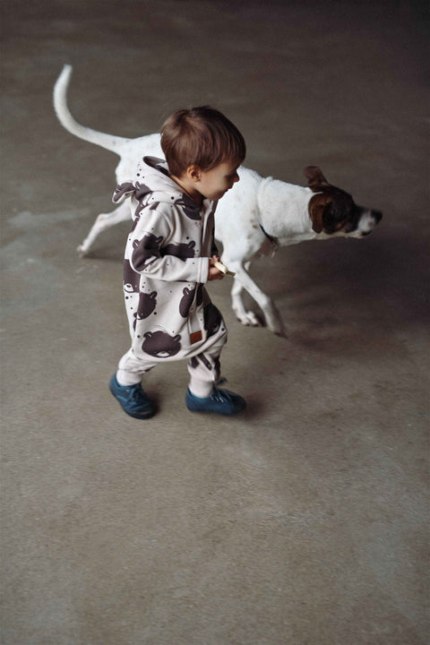 1. Child in beige bear-patterned jumpsuit with hood running alongside a dog indoors