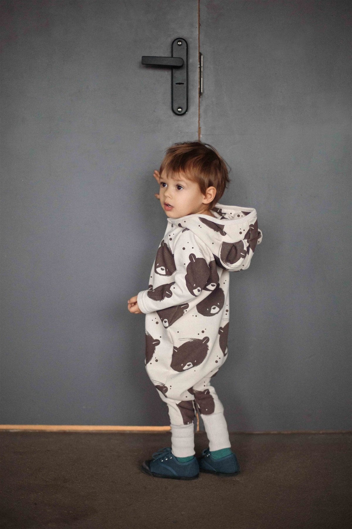 1. Toddler wearing beige bear-patterned jumpsuit with hood and ears standing by a gray door
