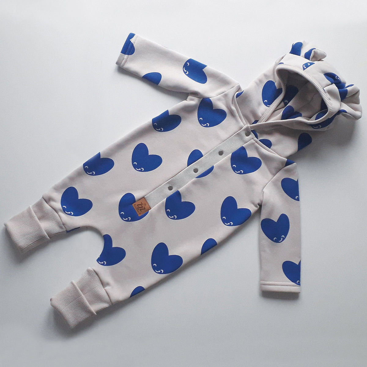 2. Eared jumpsuit for babies with blue heart print on beige fabric, shown flat with hood and foldable cuffs