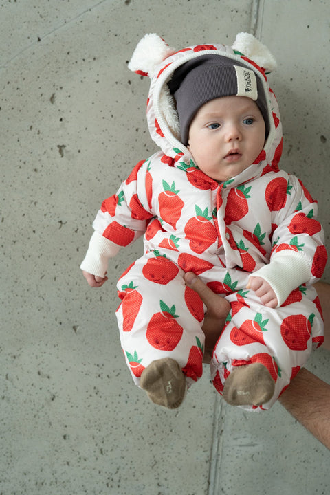 4. Baby in Zezuzulla strawberry print jumpsuit with teddy bear ears held against a concrete wall