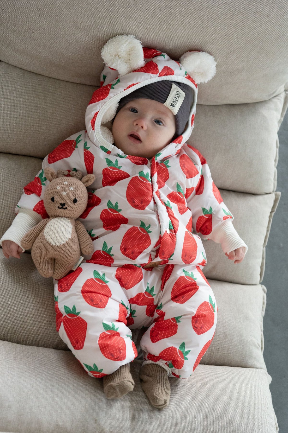 6. Baby lying on cushion in Zezuzulla strawberry print jumpsuit with teddy bear ears holding a toy