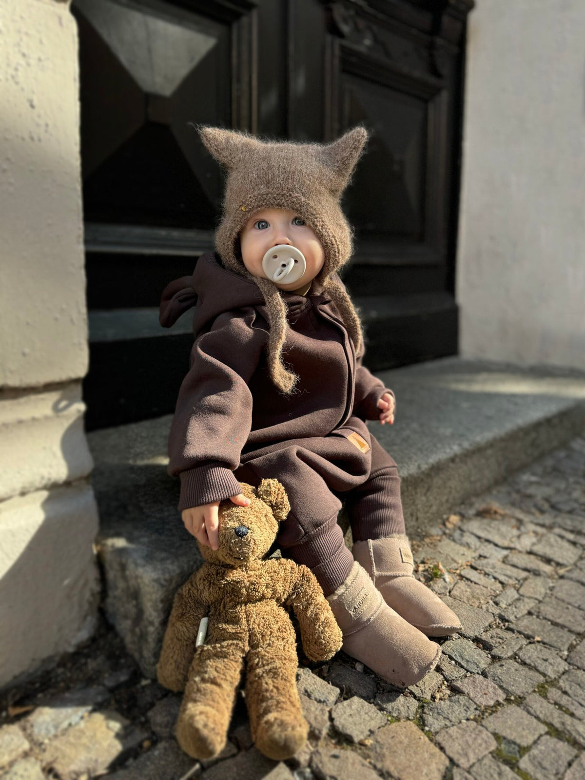 1. Baby wearing chocolate brown eared jumpsuit with hood sitting on steps holding teddy bear
