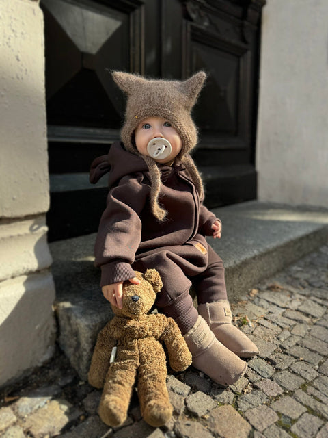 1. Baby wearing chocolate brown eared jumpsuit with hood sitting on steps holding teddy bear