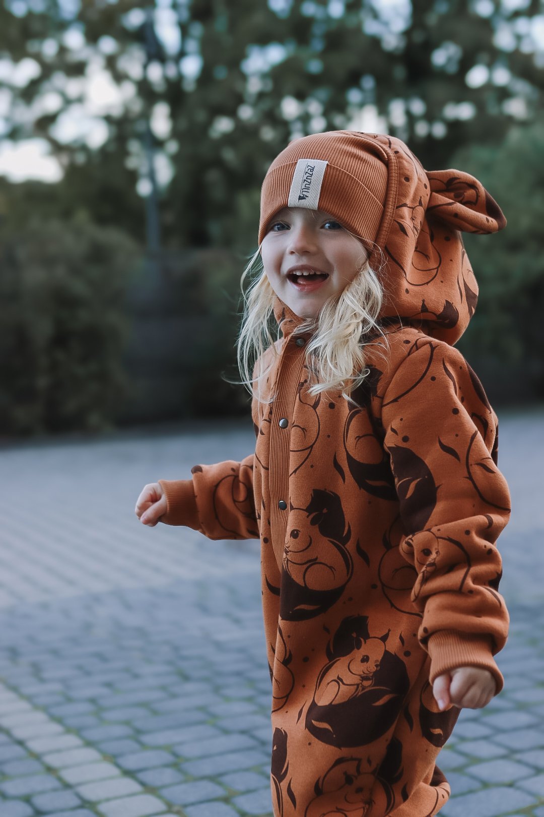 1. Child wearing cinnamon squirrel print jumpsuit with hood and ears, smiling outdoors on cobblestone path