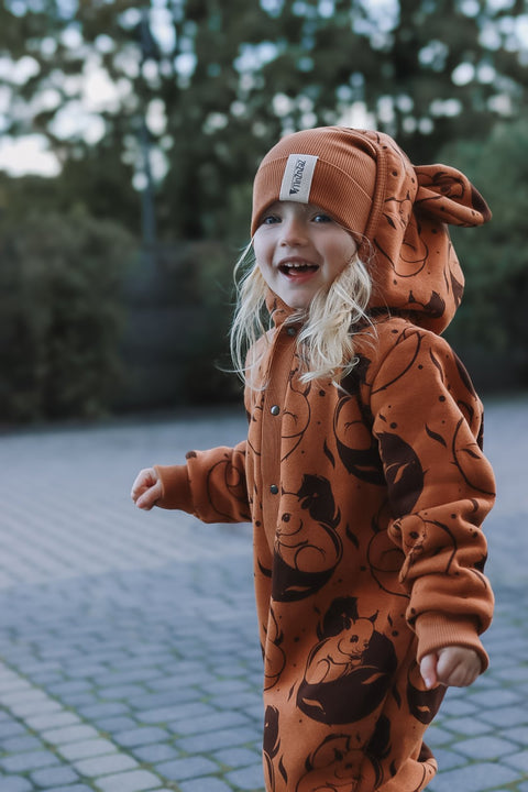 1. Child wearing cinnamon squirrel print jumpsuit with hood and ears, smiling outdoors on cobblestone path