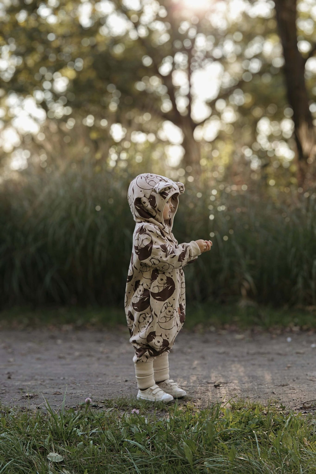 2. Child in beige squirrel print jumpsuit with hood and ears, standing on a path in a sunlit park