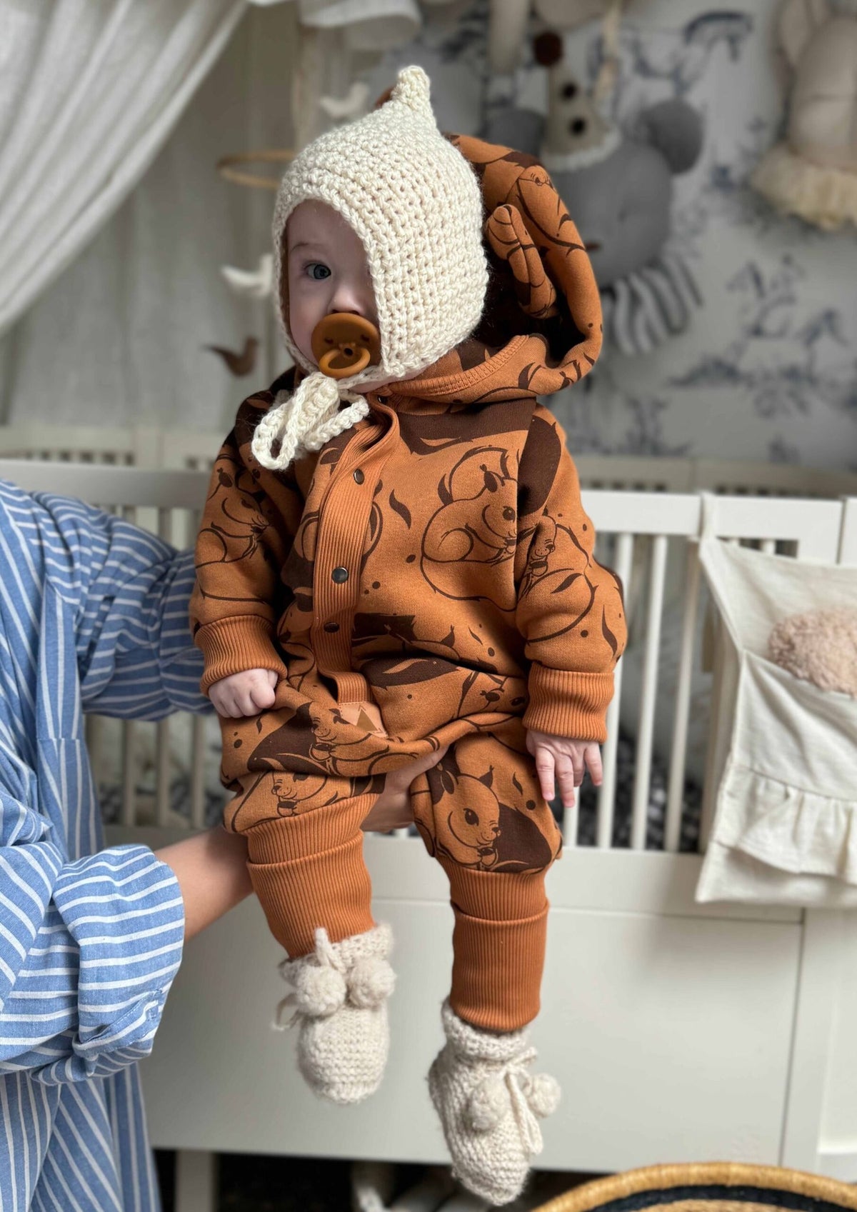 2. Baby in cinnamon squirrel jumpsuit with hood and ears, held by adult in nursery setting