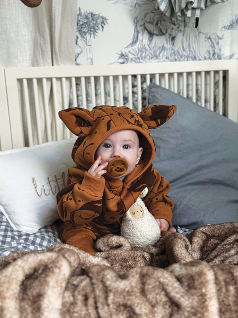 1. Baby sitting in crib wearing cinnamon squirrel jumpsuit with hood and ears, holding toy