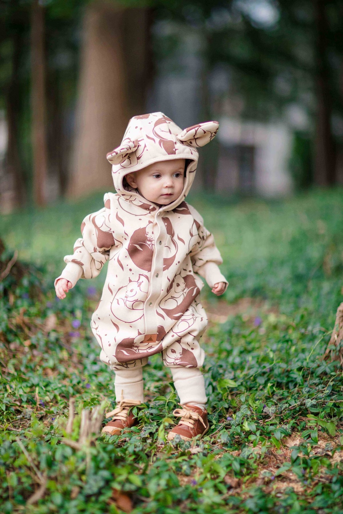 1. Baby wearing beige squirrel print jumpsuit with hood and ears, standing on grass in a park