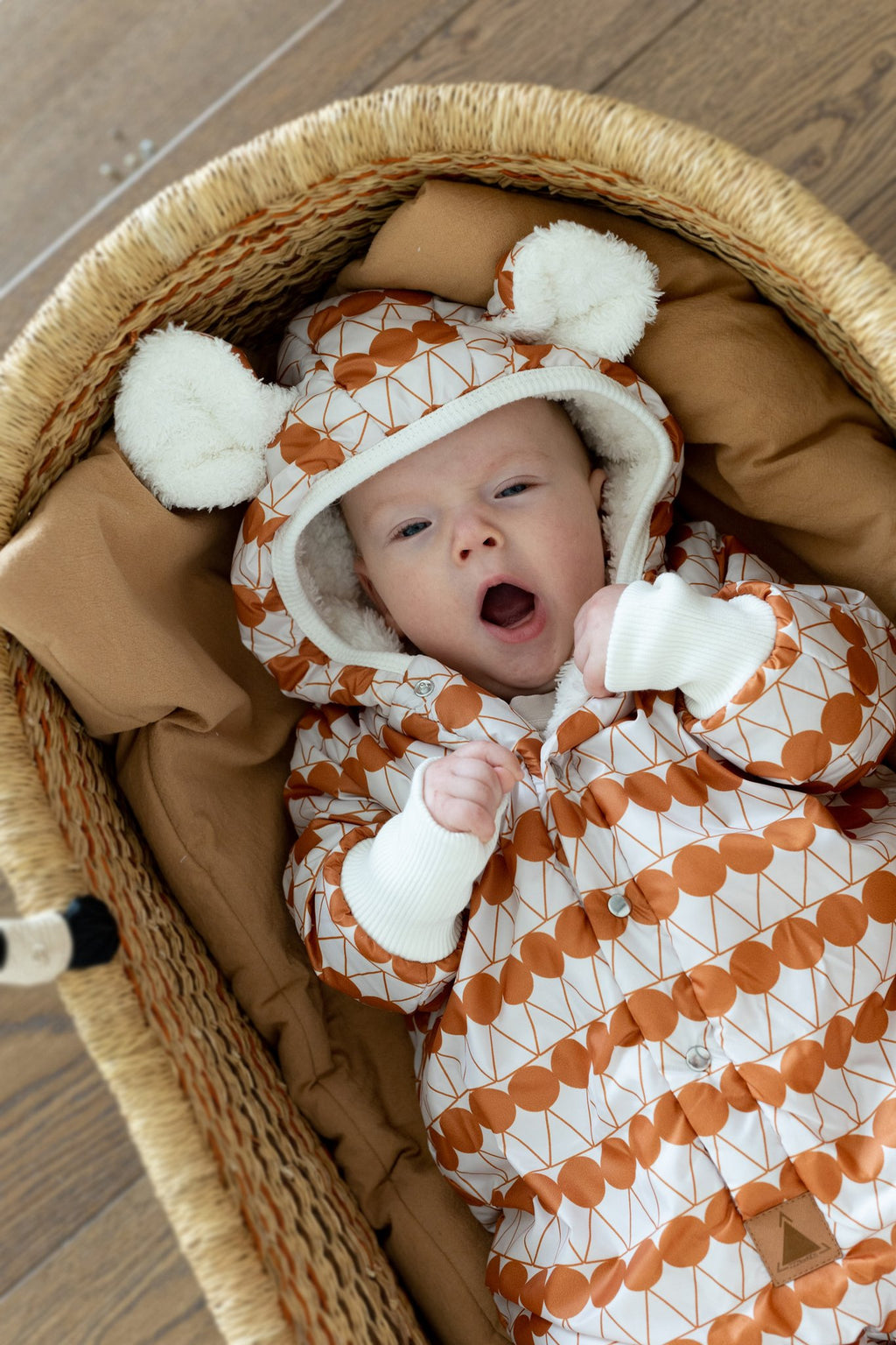 1. Baby yawning in orange dotted jumpsuit with hood and bear ears, lying in a woven basket