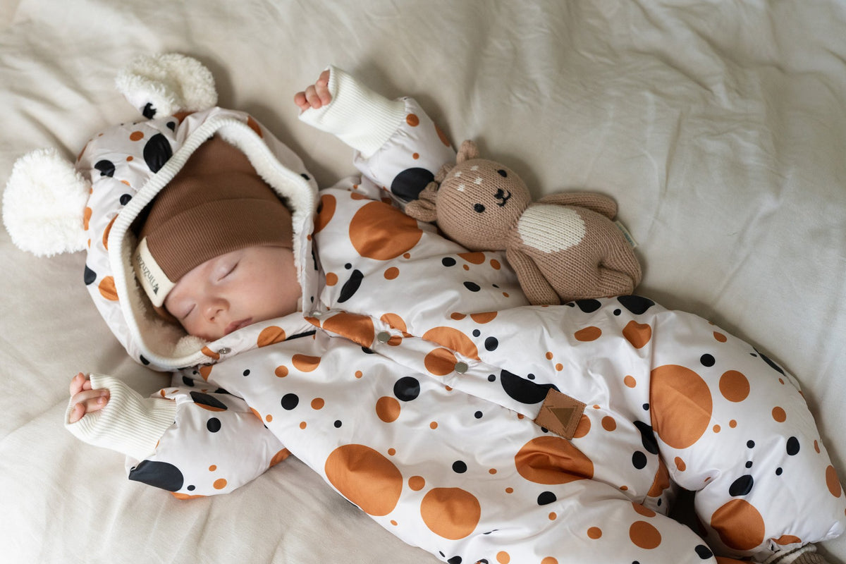 1. Baby sleeping in black and orange polka dot winter jumpsuit with teddy bear ears, lying on a bed