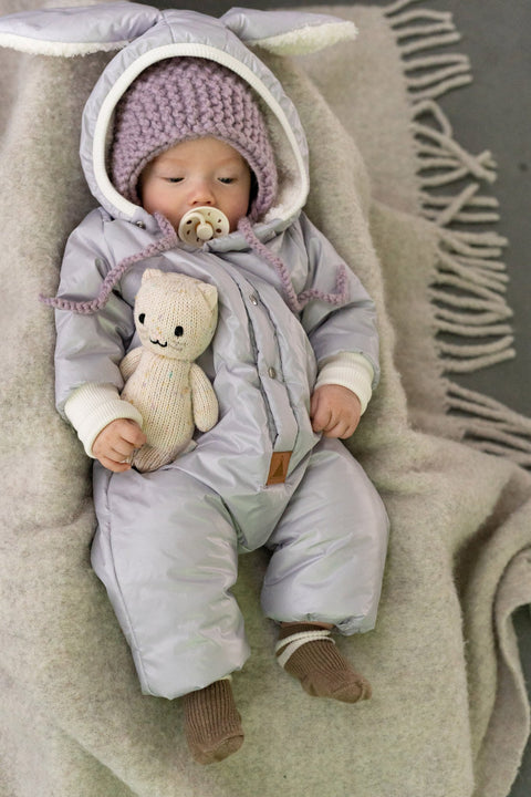 2. Baby in Zezuzulla winter jumpsuit with rabbit ears, lying on blanket, holding a knitted toy, highlighting teddy lining and foldable cuffs