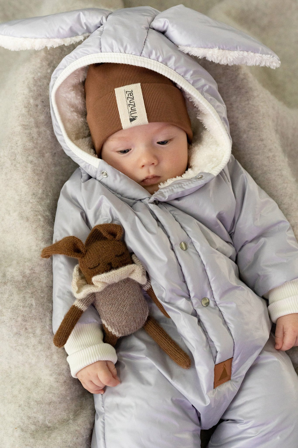 4. Baby in Zezuzulla winter jumpsuit with rabbit ears, wearing a brown hat, holding a stuffed toy, showcasing cozy teddy lining and waterproof fabric