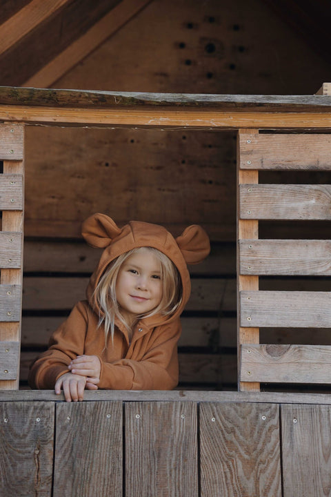 1. Child wearing cinnamon eared jumpsuit with hood, smiling while leaning on wooden playhouse