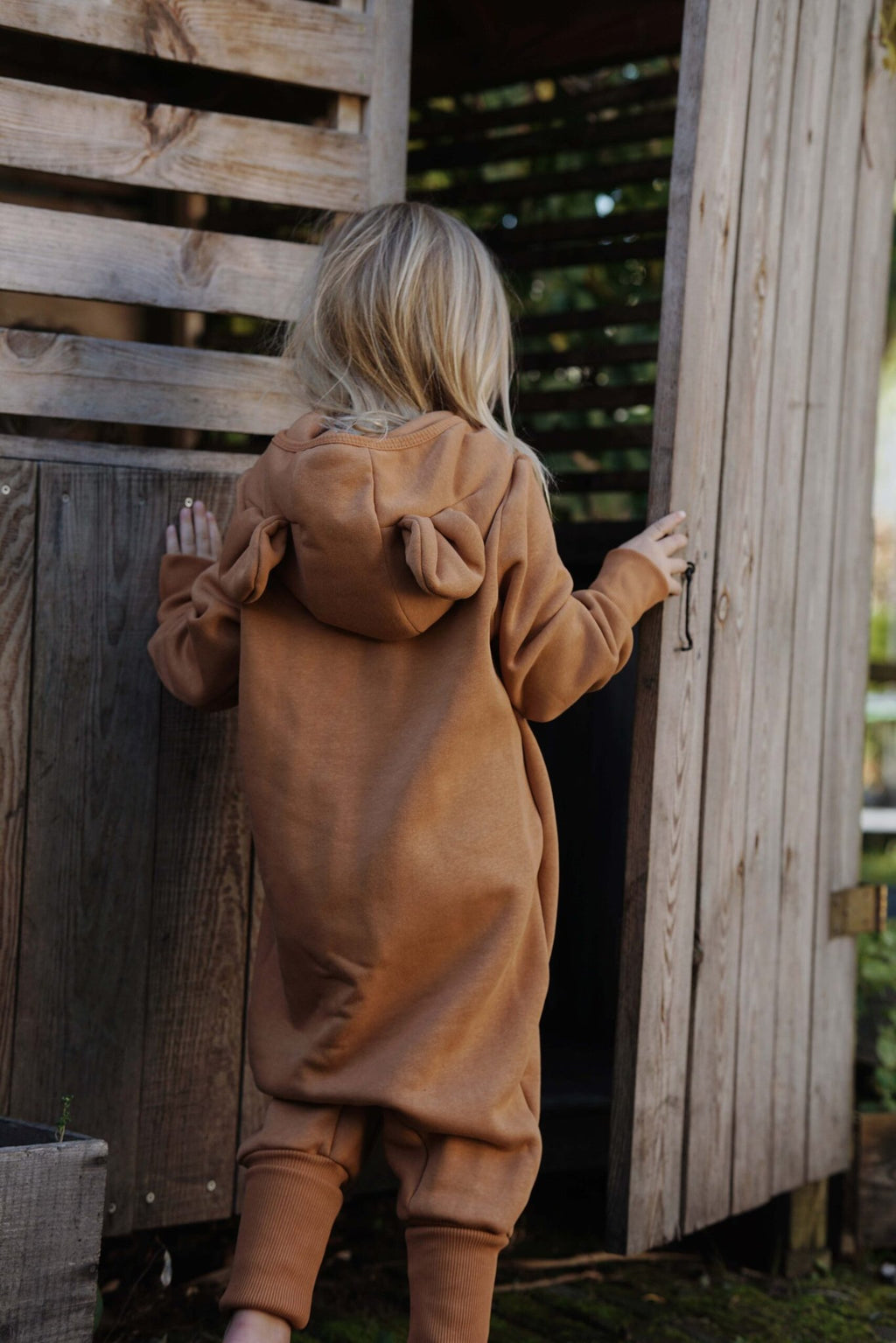 1. Child in cinnamon eared jumpsuit with hood, walking into wooden playhouse, shown from back