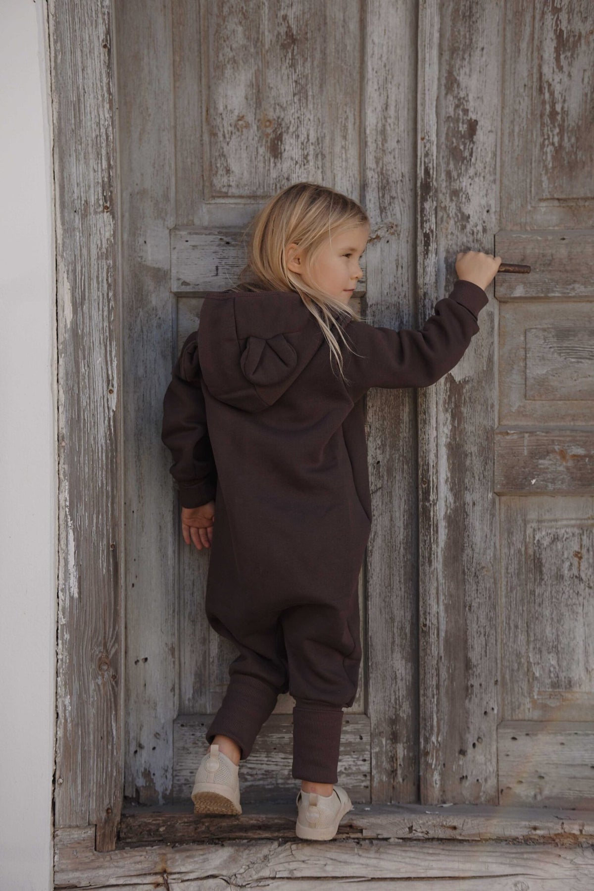 4. Back view of child in chocolate brown eared jumpsuit with hood against wooden door