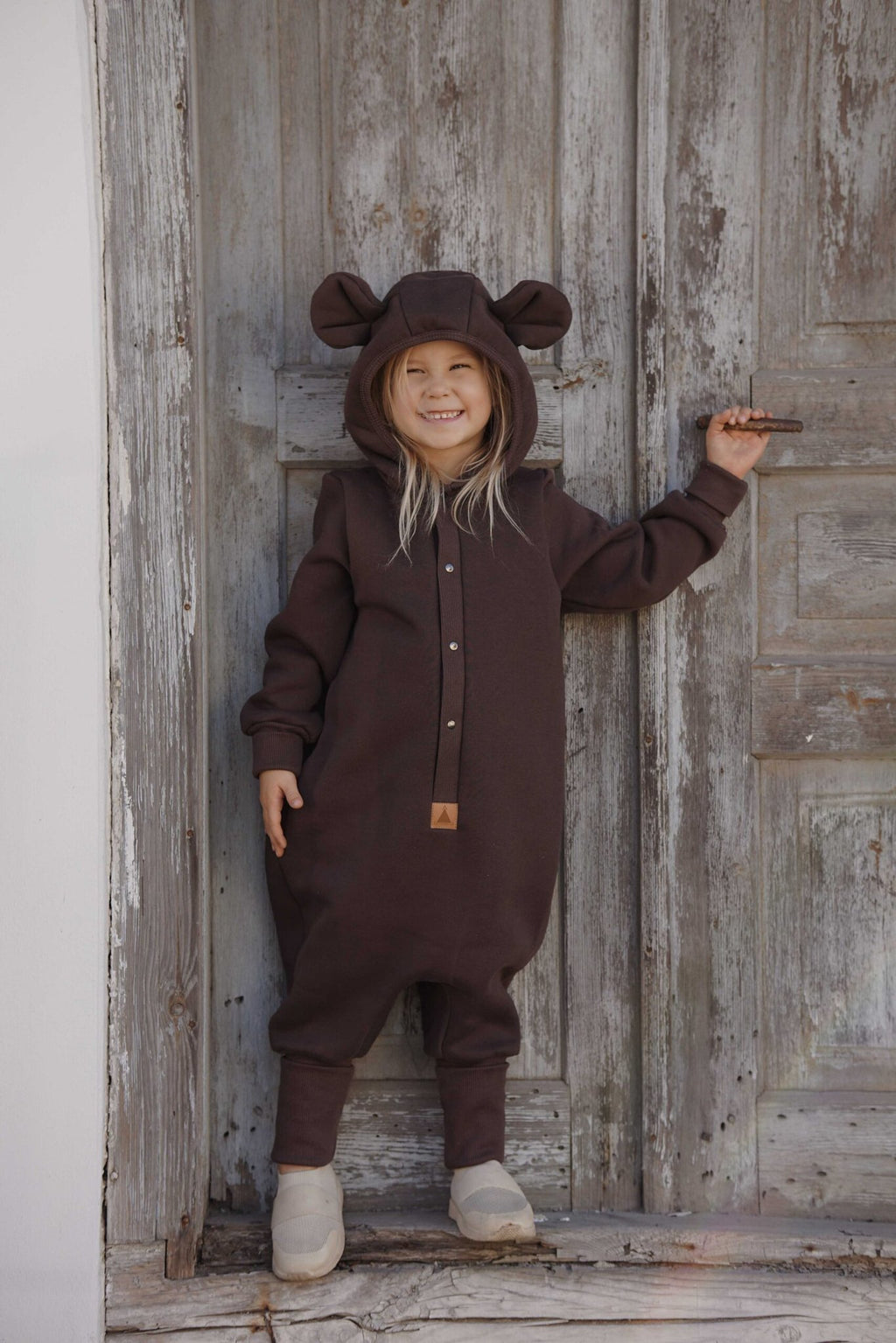 3. Child wearing chocolate brown eared jumpsuit with hood standing against rustic wooden door