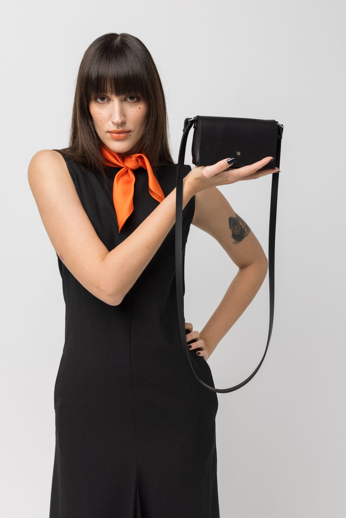 1. Woman holding PYKOK KORA minibag in black leather, styled with orange scarf and black dress in studio setting