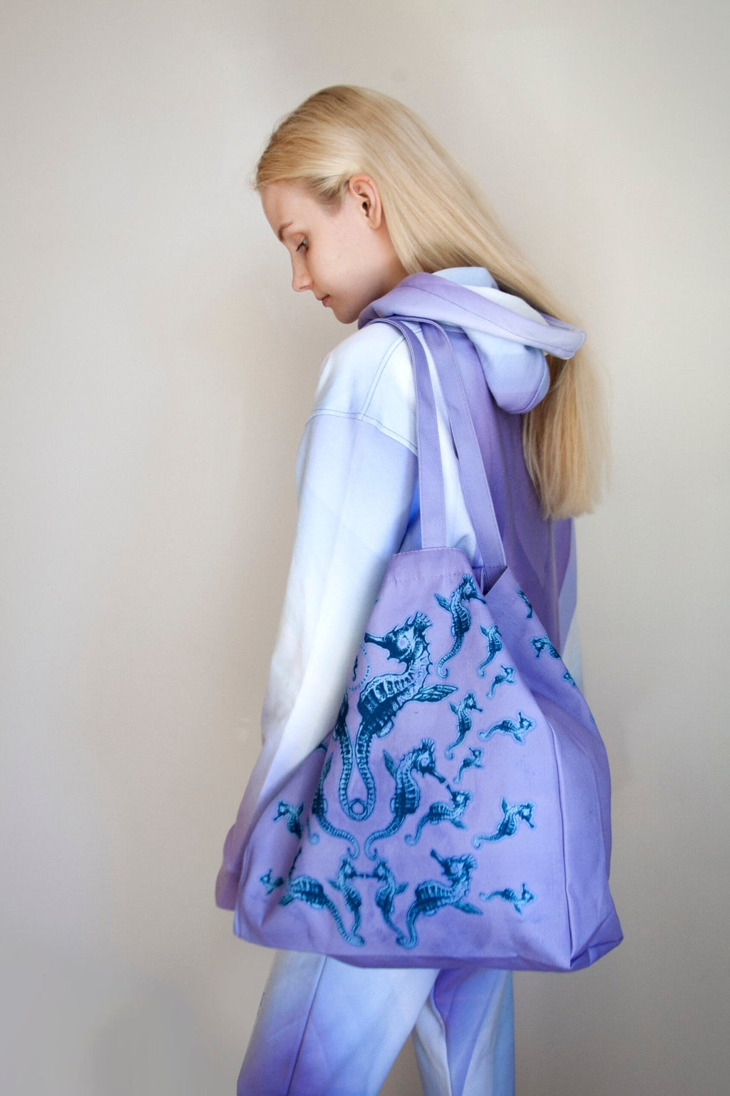 2. Woman in pastel hoodie carrying a Tanel Veenre Mirage tote bag with blue seahorse design, highlighting spacious size and long handles, in a neutral indoor setting