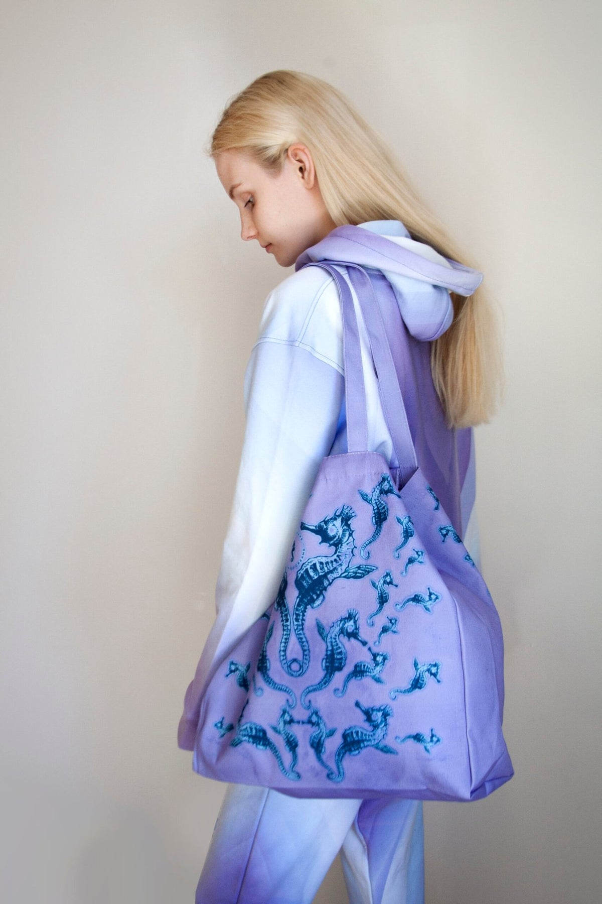 2. Woman in pastel hoodie carrying a Tanel Veenre Mirage tote bag with blue seahorse design, highlighting spacious size and long handles, in a neutral indoor setting