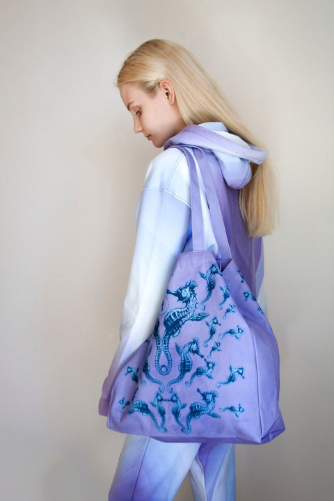 2. Woman in pastel hoodie carrying a Tanel Veenre Mirage tote bag with blue seahorse design, highlighting spacious size and long handles, in a neutral indoor setting