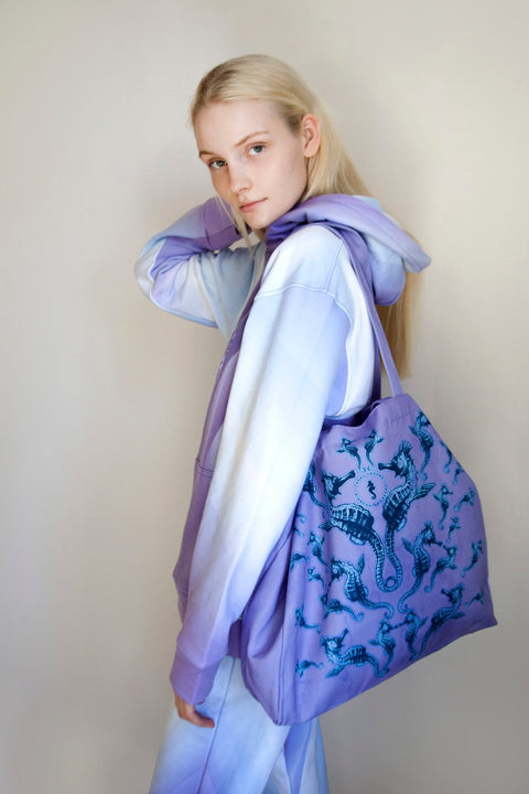 1. Woman wearing a pastel hoodie holding a Tanel Veenre Mirage tote bag with blue seahorse print, showcasing long handles, in a neutral indoor setting