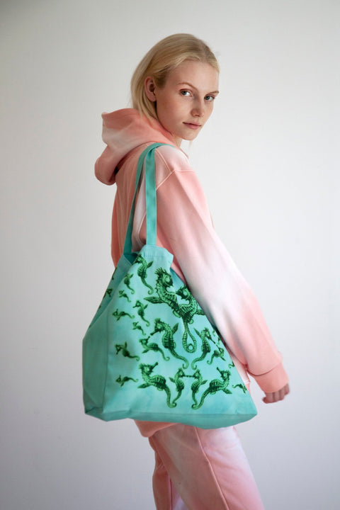 2. Woman in pink and white hoodie carrying Tanel Veenre Mirage tote bag featuring green seahorse pattern