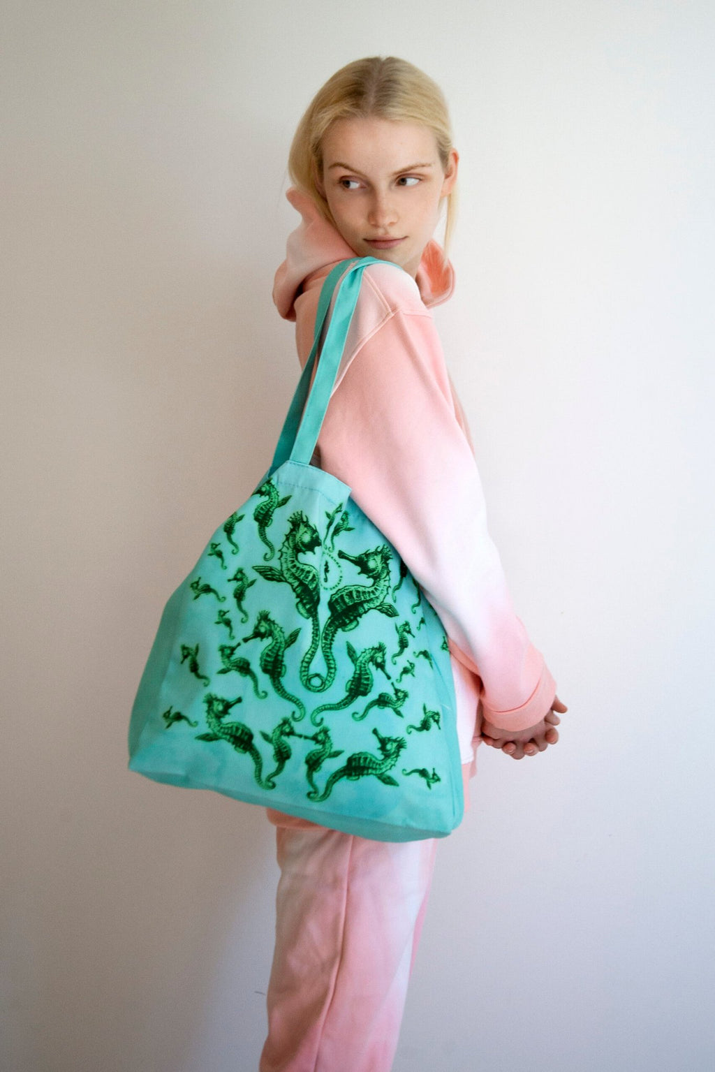 1. Woman wearing pink and white hoodie holding Tanel Veenre Mirage tote bag with green seahorse design