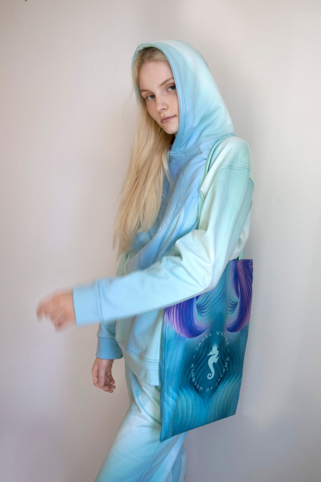 3. Woman in a pastel blue hoodie with a Tanel Veenre Mirage tote bag featuring a seahorse design, looking down against a plain background