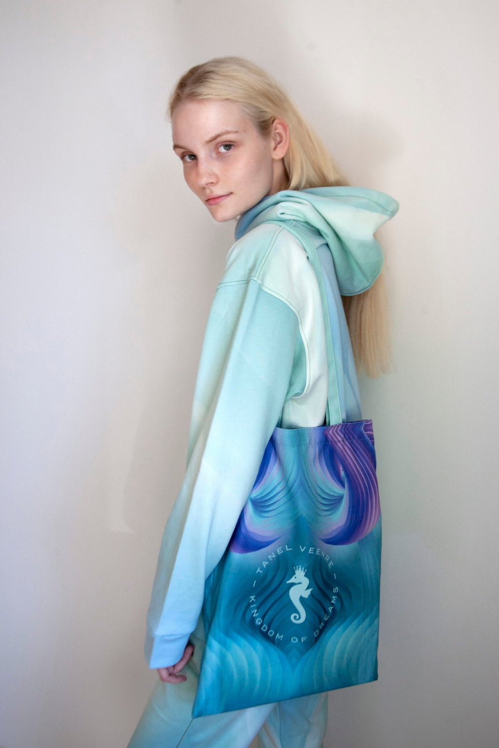 1. Woman wearing a pastel blue hoodie carrying a Tanel Veenre Mirage tote bag with a seahorse design, standing against a plain background