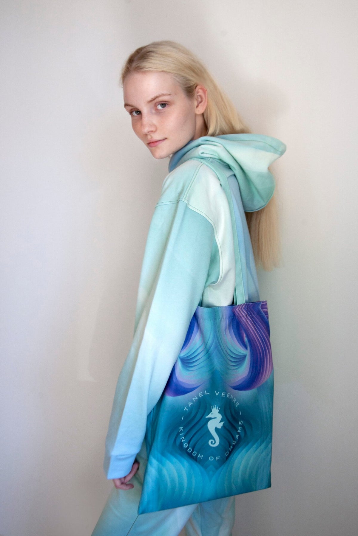 1. Woman wearing a pastel blue hoodie carrying a Tanel Veenre Mirage tote bag with a seahorse design, standing against a plain background
