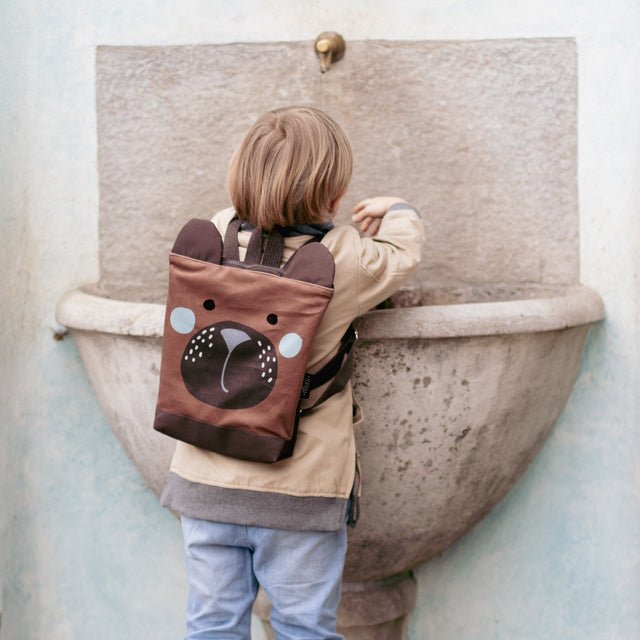 1. Child wearing Muni Green Bear Backpack at a stone fountain, showcasing playful design
