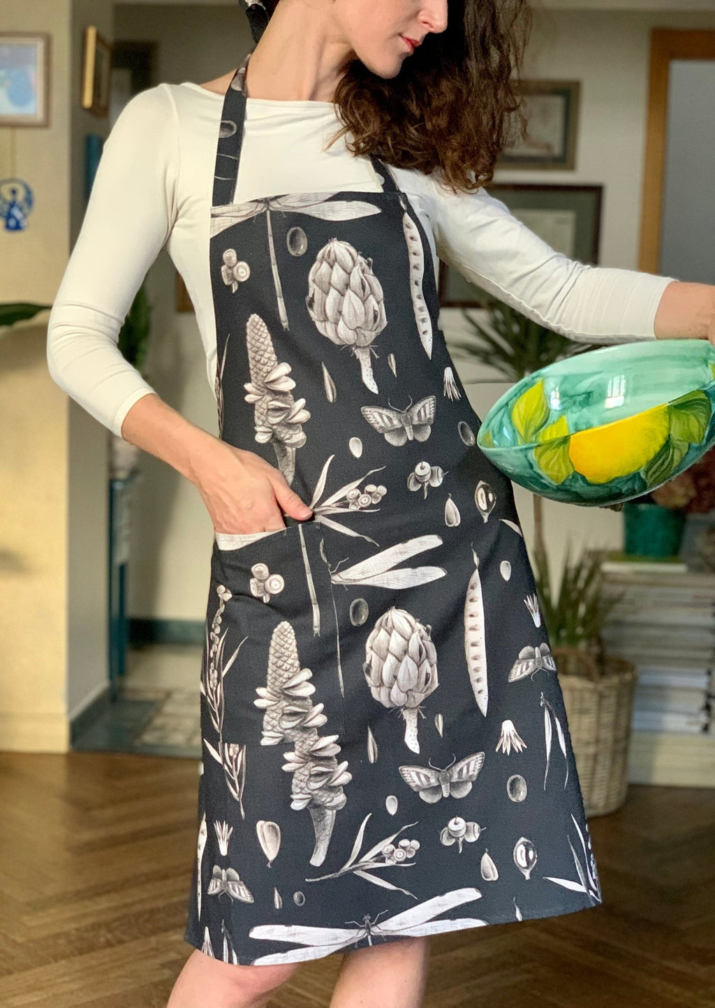1. Woman wearing Hortensias Home Seeds Carbon apron with botanical print, holding a bowl in a cozy kitchen setting