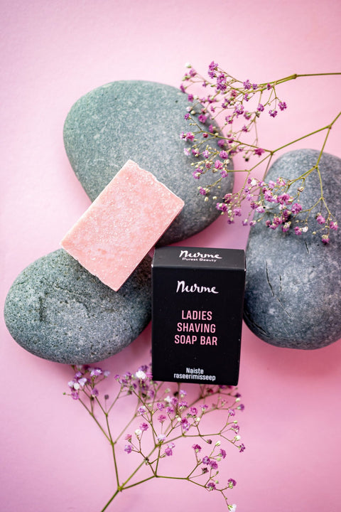 4. Flat lay of Nurme Ladies Shaving Soap Bar with pink soap and black packaging on pink background with stones and flowers