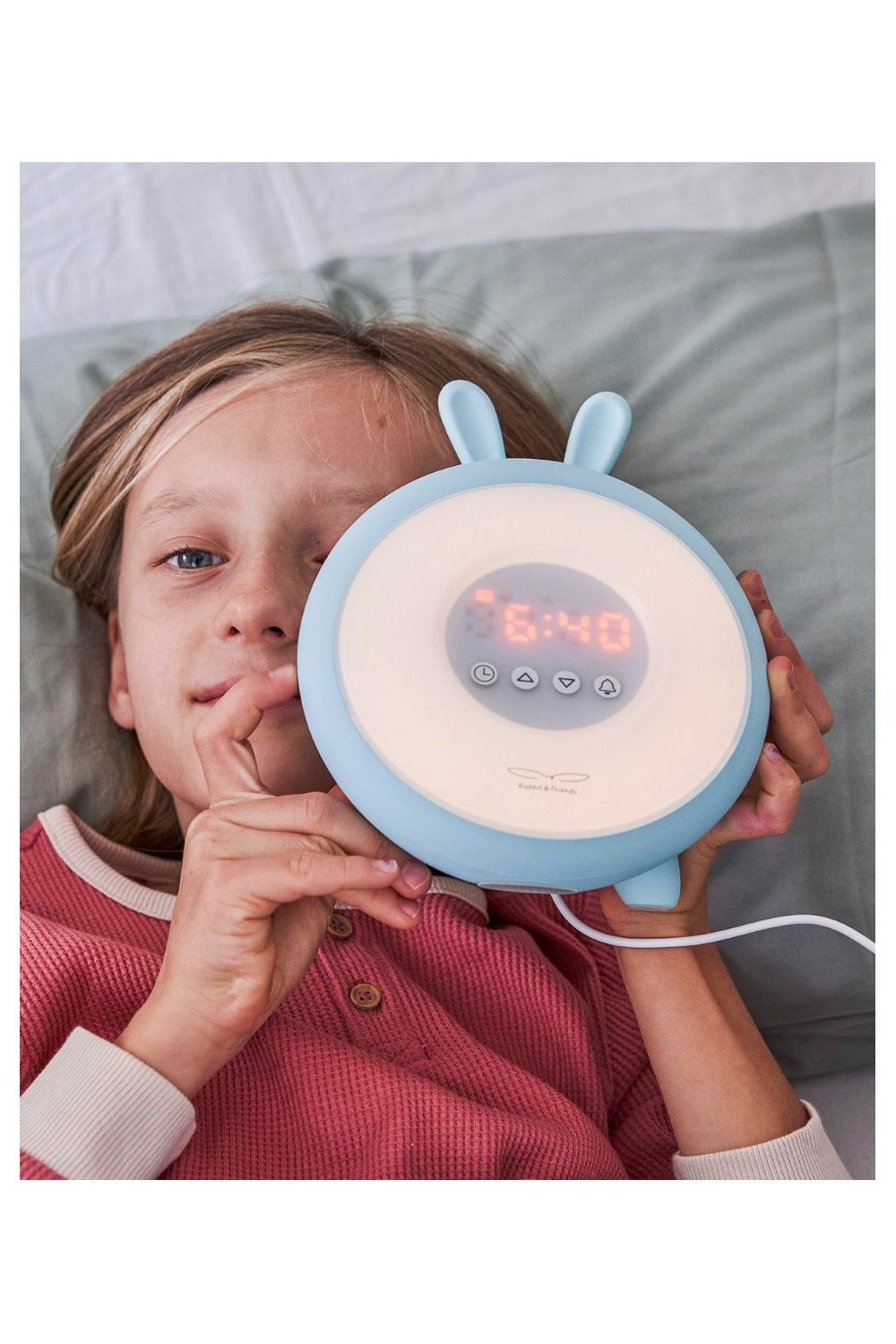 2. Child holding blue Rabbit & Friends alarm lamp with ears, showing digital time, lying on bed