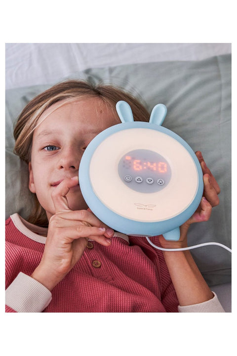 2. Child holding blue Rabbit & Friends alarm lamp with ears, showing digital time, lying on bed