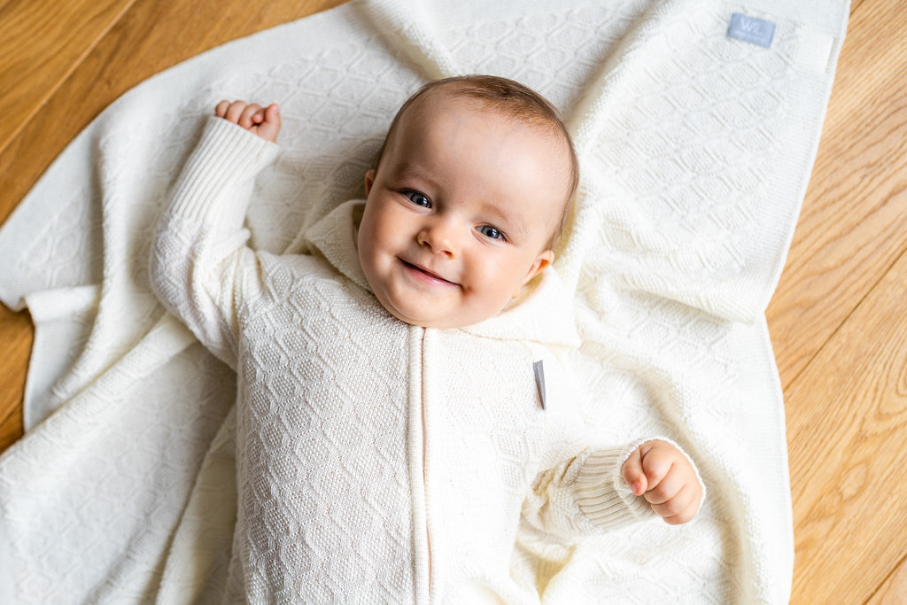 4. Smiling baby in white merino wool romper on matching blanket on wooden floor
