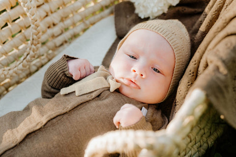 4. Baby in brown merino wool romper and bonnet lying in a basket with a braided blanket