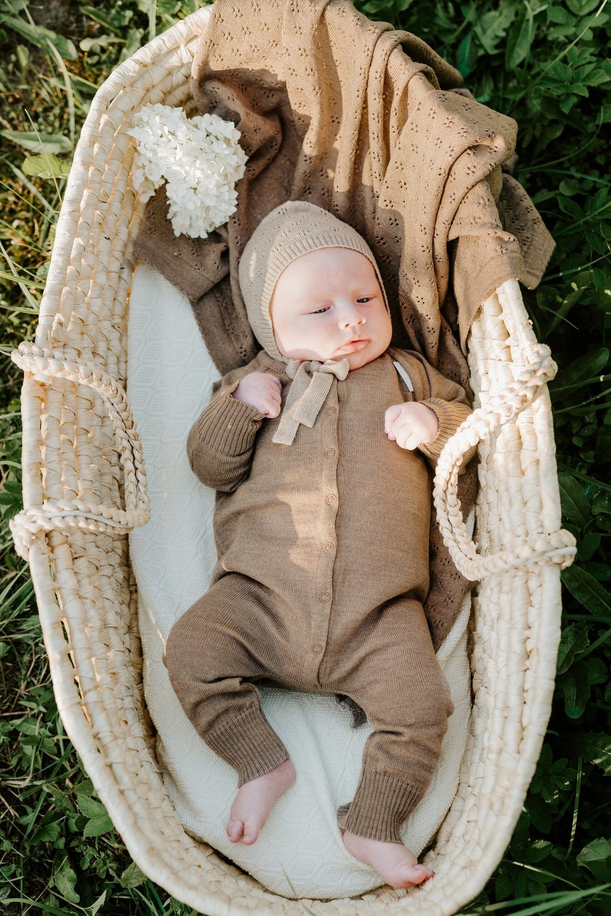 1. Baby wearing brown merino wool romper and bonnet lying in a woven basket with a braided blanket outdoors