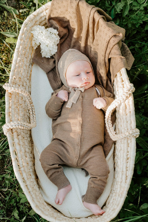 1. Baby wearing brown merino wool romper and bonnet lying in a woven basket with a braided blanket outdoors