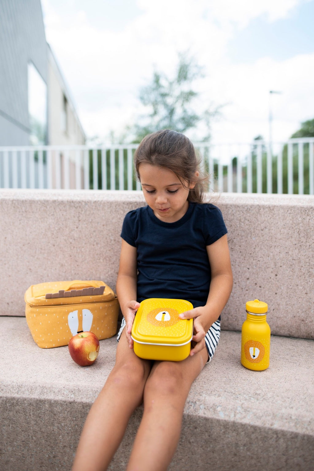 1. Young girl sitting on steps holding yellow Mr. Lion lunch box with matching bottle and larger lunch box nearby
