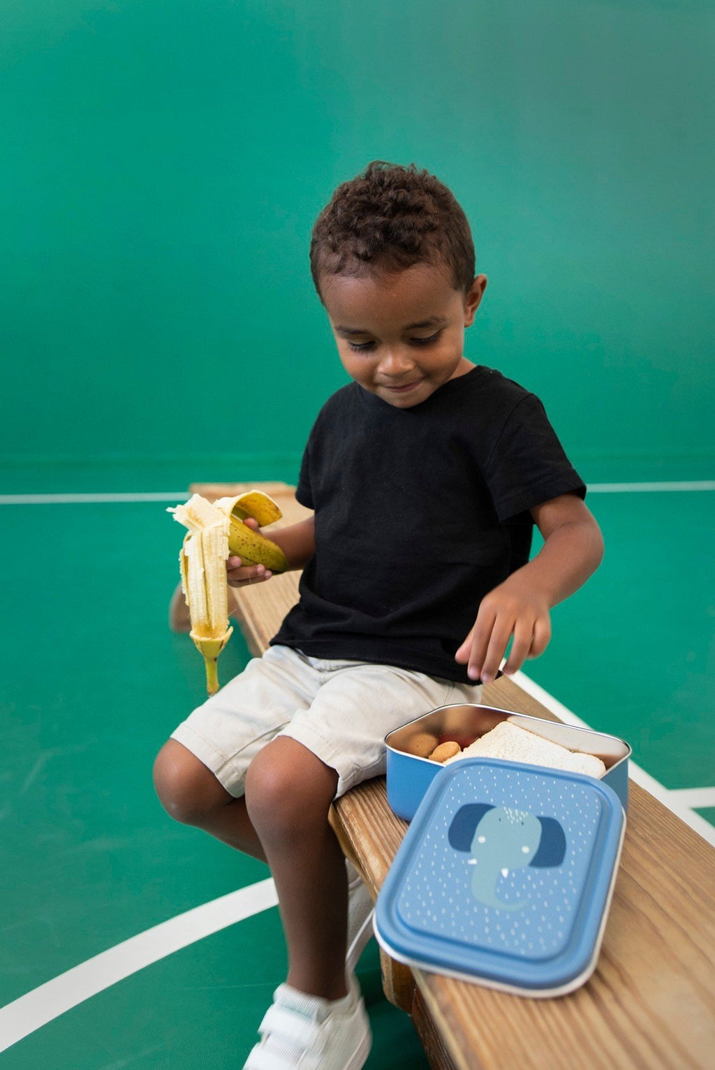 1. Child sitting on a bench with Trixie Baby Mrs. Elephant lunch box, enjoying a banana, in a playful setting