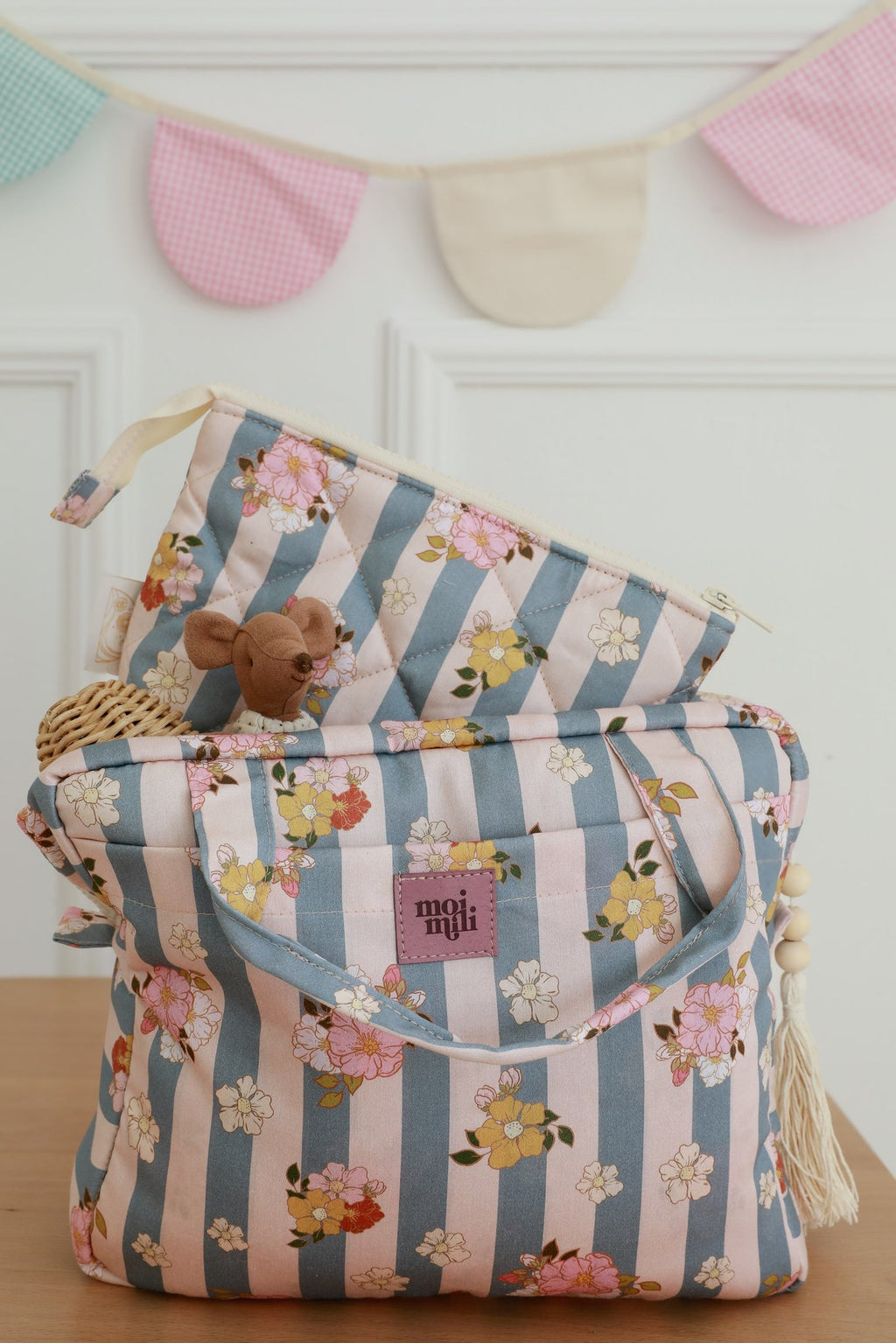1. Two blue striped makeup bags with floral print on wooden table, featuring Moi Mili logo, with small toy and decorative bunting in background