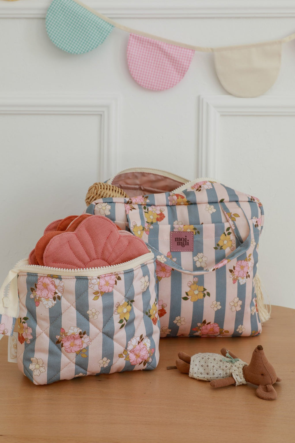 5. Two blue striped floral makeup bags with Moi Mili logo on wooden table, small toy and decorative bunting in background