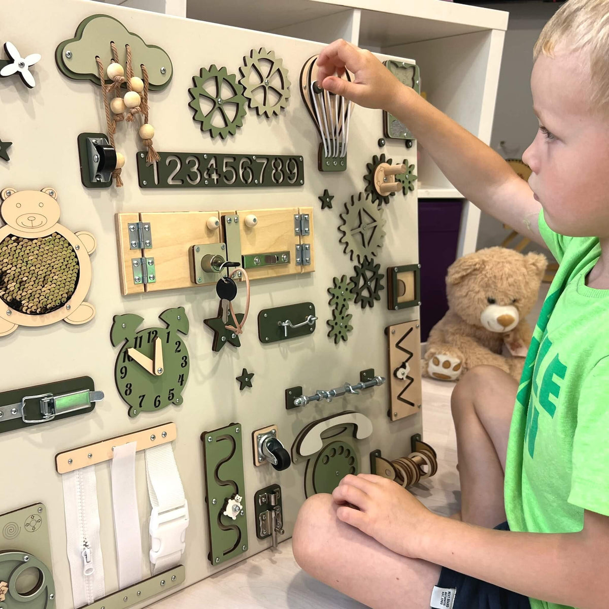 5. Child interacting with large Montessori busy board featuring green and beige elements, in a playroom setting.
