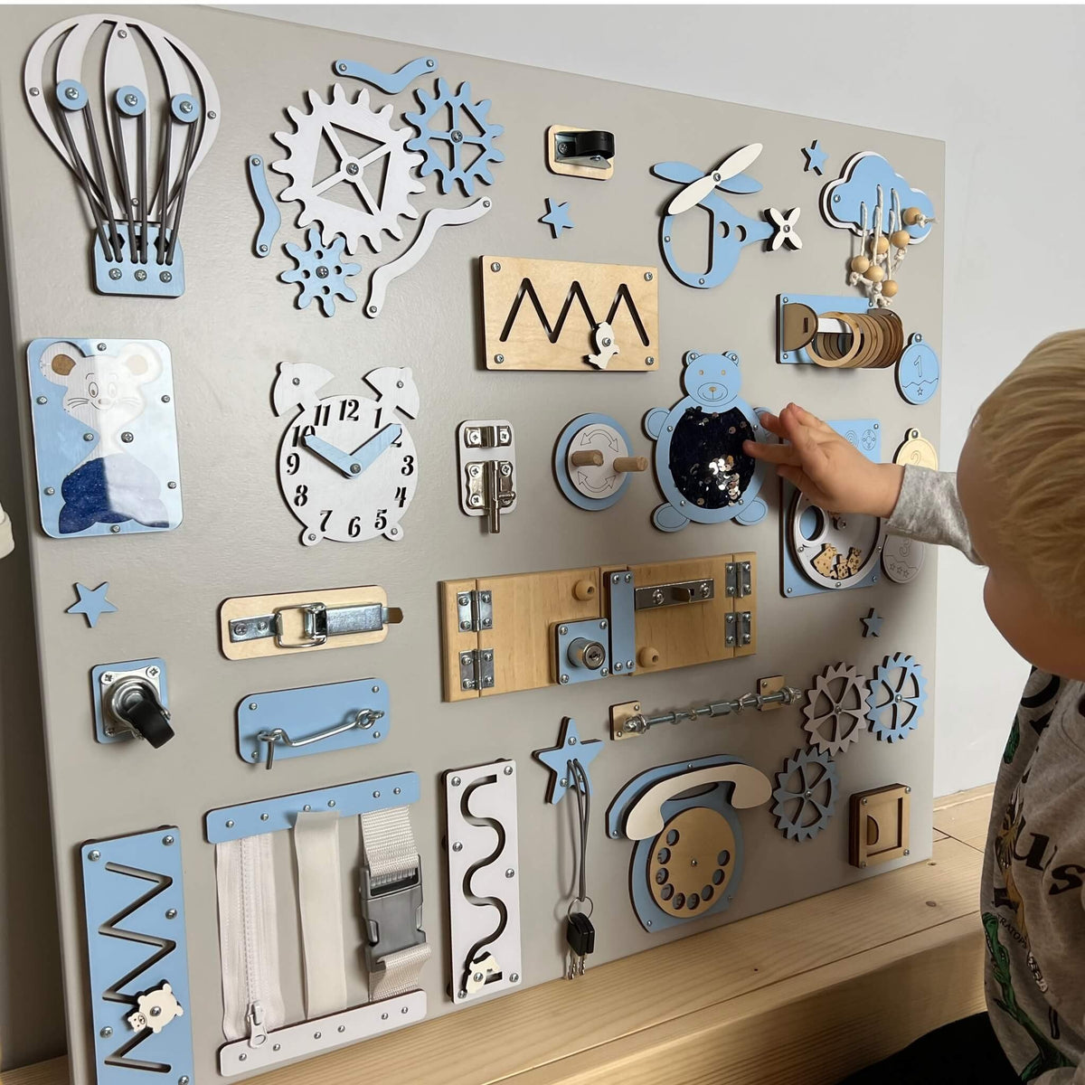 5. Child playing with large wall busy board featuring blue and white elements like gears, locks, and a telephone on a grey background