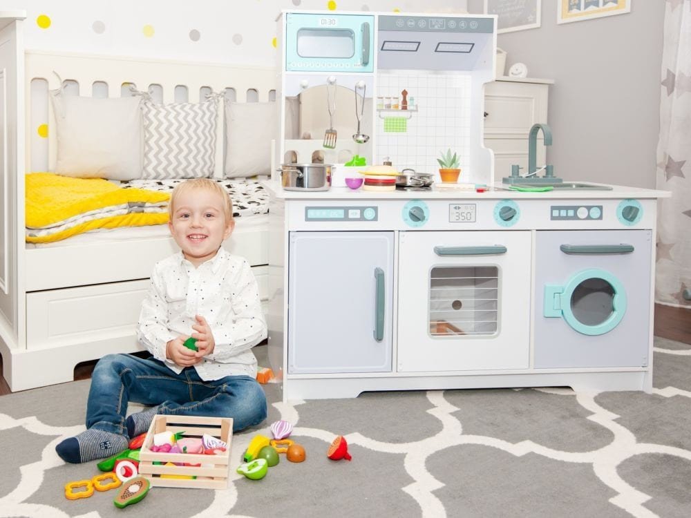 2. Smiling child sitting on carpet with Angelove wooden play kitchen in bedroom setting, showing oven, microwave, and washing machine