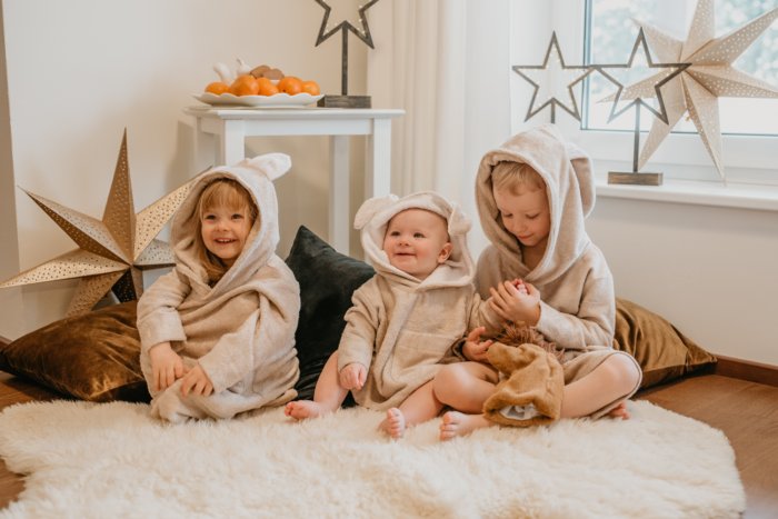 1. Three toddlers wearing RÄTT long-sleeve poncho TEDDY in beige, sitting on a fluffy rug in a cozy room setting