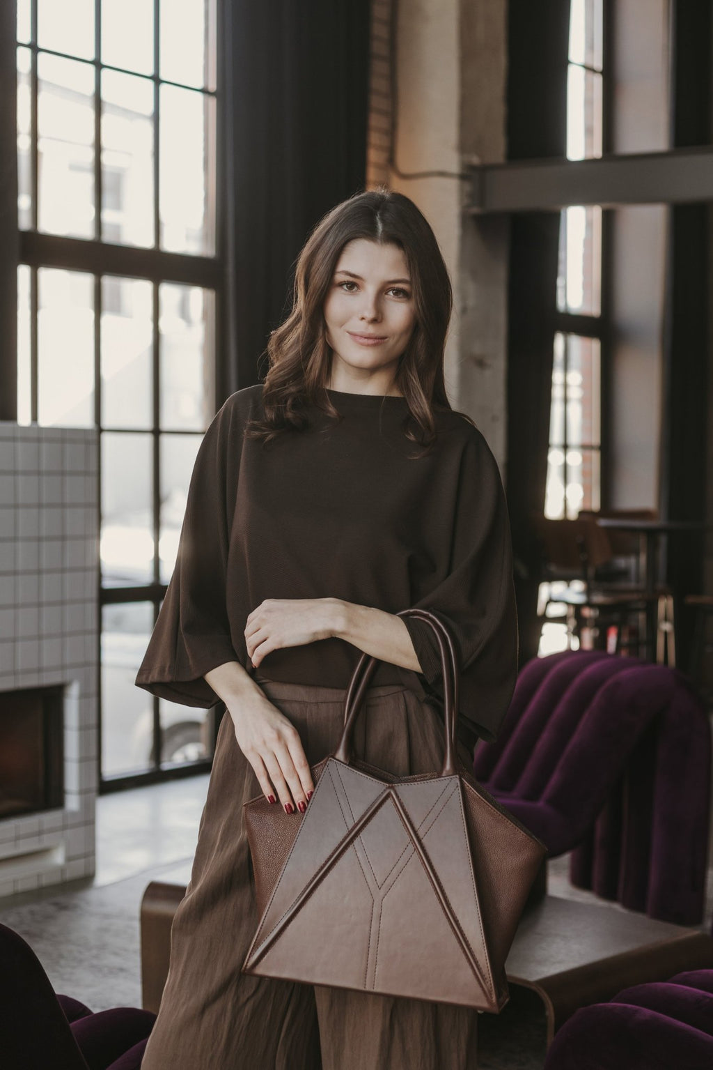 2. Woman with Gerda Retter Design Leather Handbag Nova in dark brown, standing in a modern interior with large windows